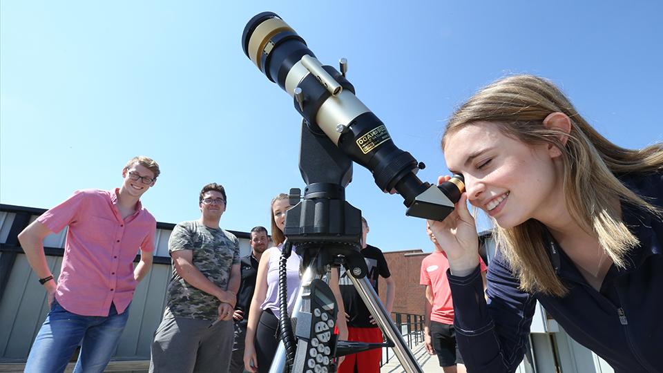 in the foreground, a student smiles while looking through the eyepiece of a telescope pointed toward the blue sky. In the background six other students stand in a line behind her and smile toward the camera.